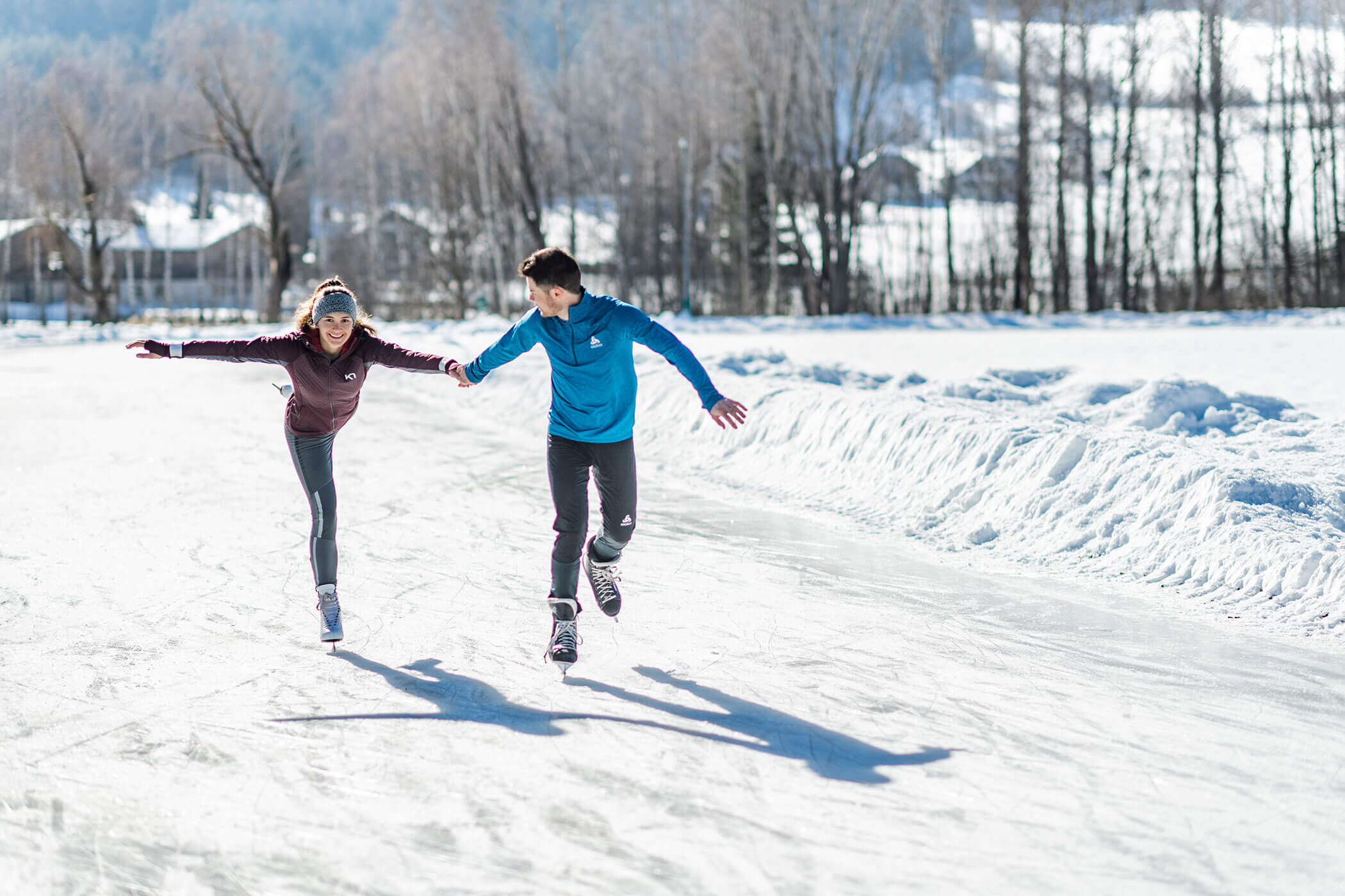 Zwei Menschen, die an einem sonnigen Wintertag im Freien Schlittschuh laufen, Händchen haltend, mit Schnee und Bäumen im Hintergrund. - Hotel Oberwiesen
