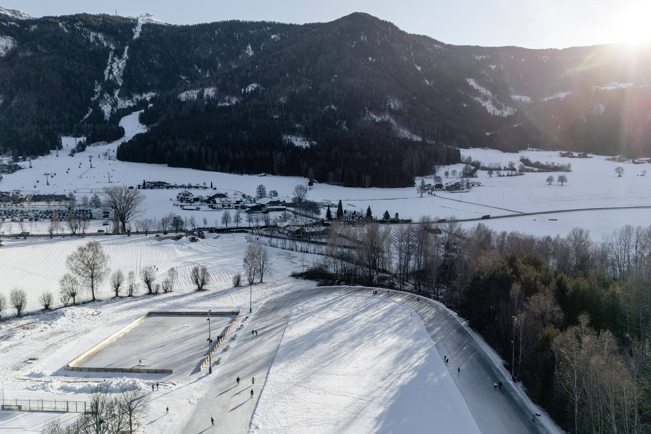 Luftaufnahme von Menschen, die auf einer Freiluft-Eisbahn in einer verschneiten Berglandschaft unter strahlend blauem Himmel Eislaufen. - Hotel Oberwiesen