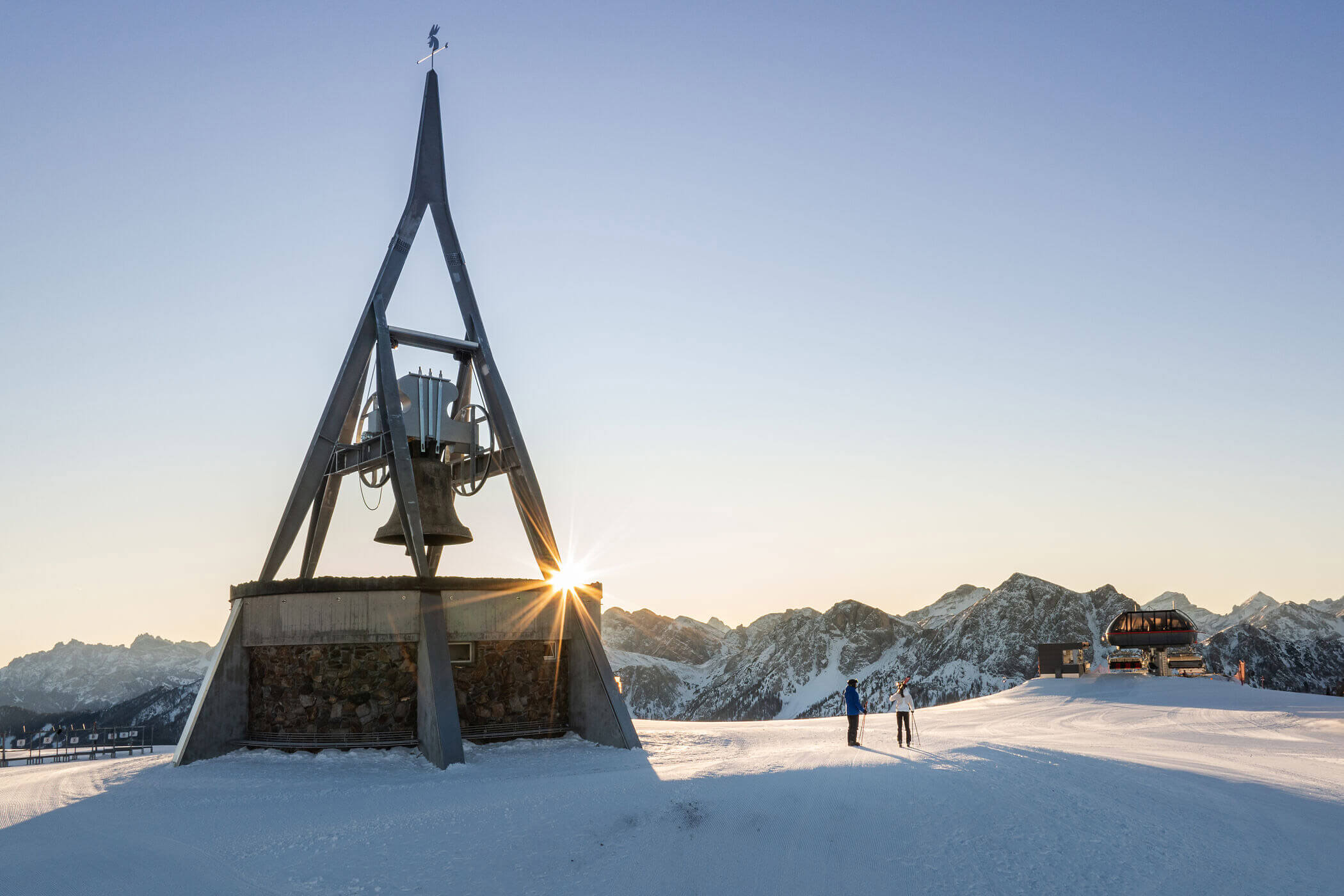 Dreieckiges Metalldenkmal auf einem schneebedeckten Berg bei Sonnenaufgang, mit zwei Skifahrern und einem Chalet in der Ferne, dahinter die Berggipfel. - Hotel Oberwiesen