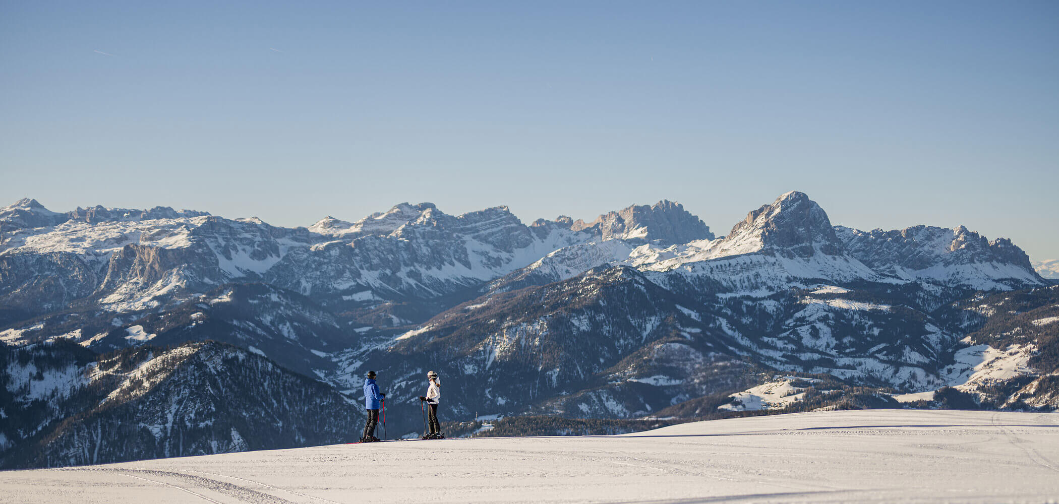 Zwei Menschen stehen auf einem verschneiten Hang, im Hintergrund ragen hohe, schroffe Berge empor, über ihnen erstreckt sich ein klarer Himmel. - Hotel Oberwiesen