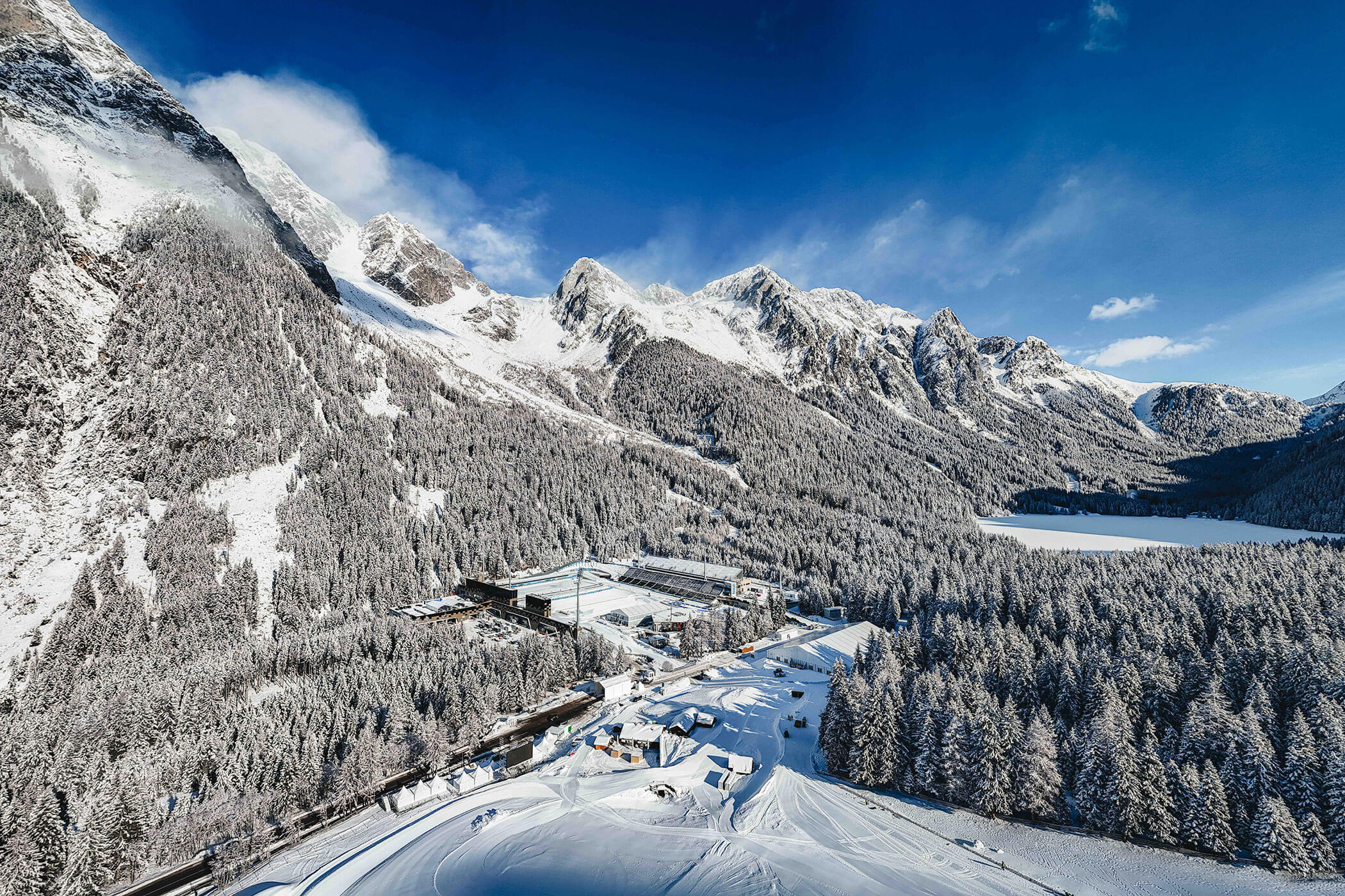 Verschneite Berge, dichter Wald und Gebäude umgeben einen zugefrorenen See unter einem strahlend blauen Himmel. - Hotel Oberwiesen
