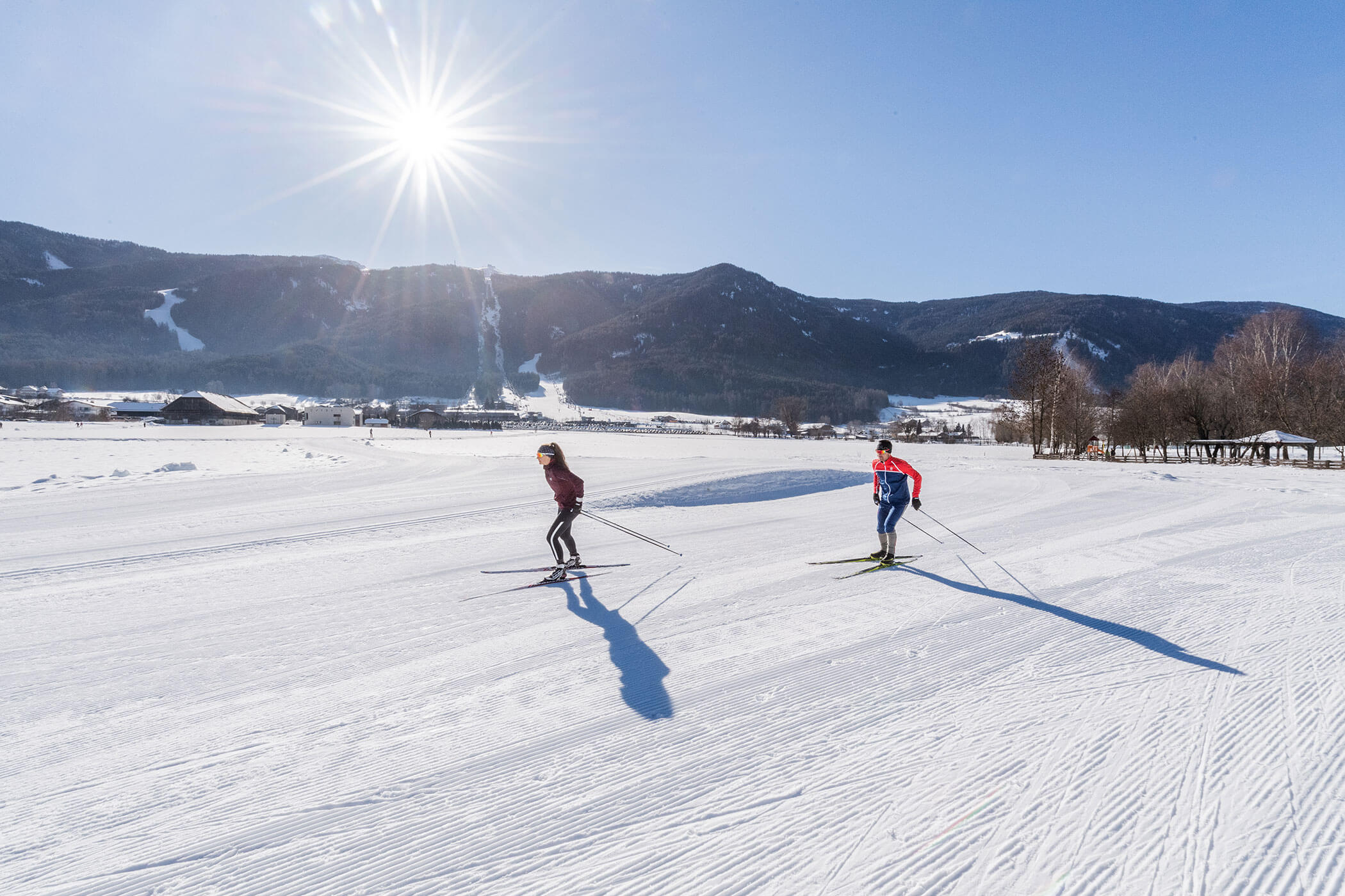 Zwei Menschen fahren auf einem sonnigen, verschneiten Feld Langlauf, im Hintergrund sind Berge zu sehen. - Hotel Oberwiesen