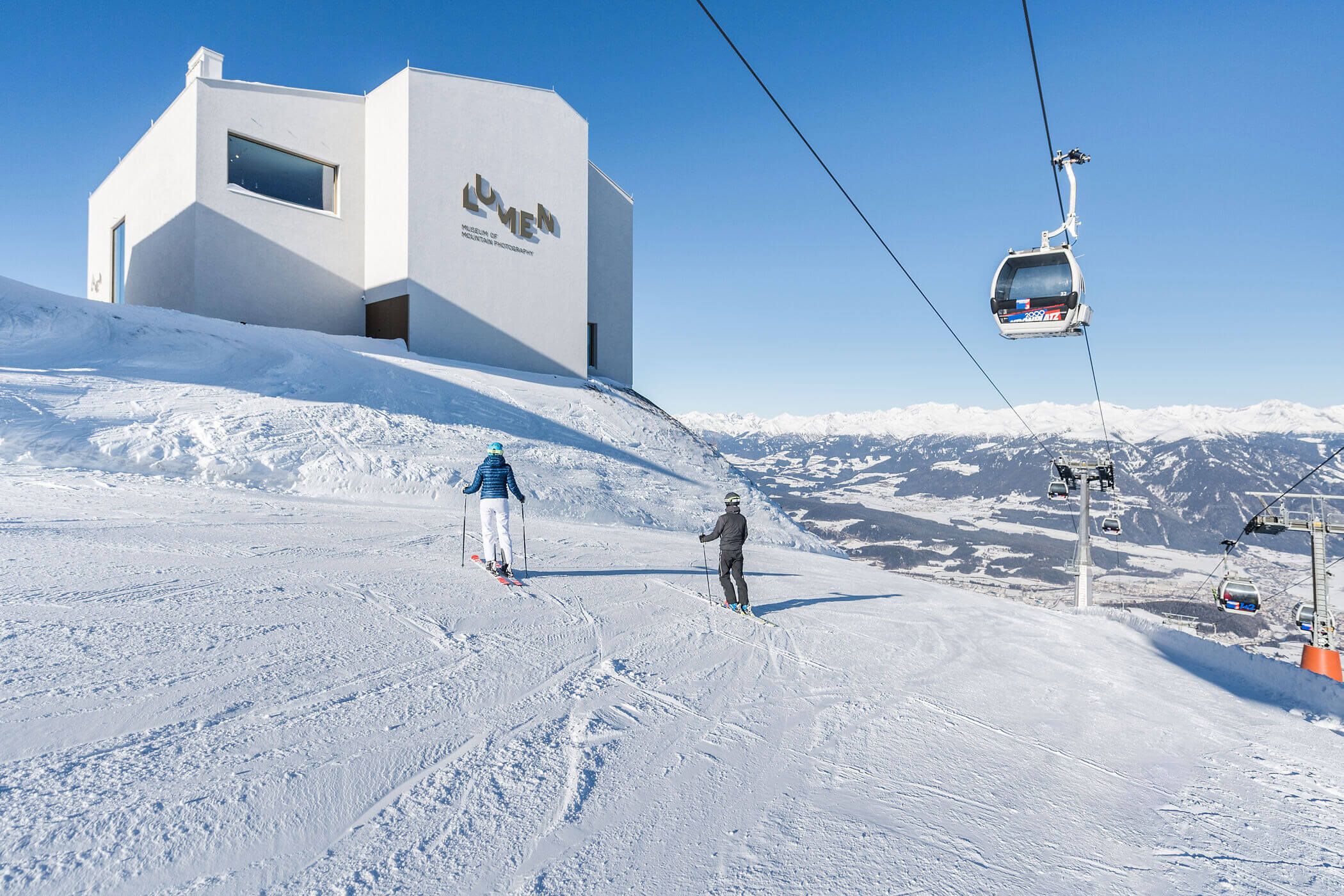 Zwei Skifahrer in der Nähe eines modernen weißen Gebäudes und eines Skilifts auf einem verschneiten Berg vor einem strahlend blauen Himmel. - Hotel Oberwiesen