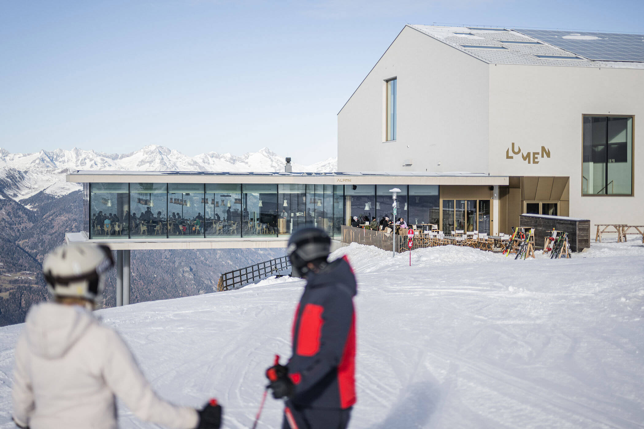 Zwei Skifahrer stehen im Schnee vor einem modernen Bergrestaurant mit großen Glasfenstern. - Hotel Oberwiesen