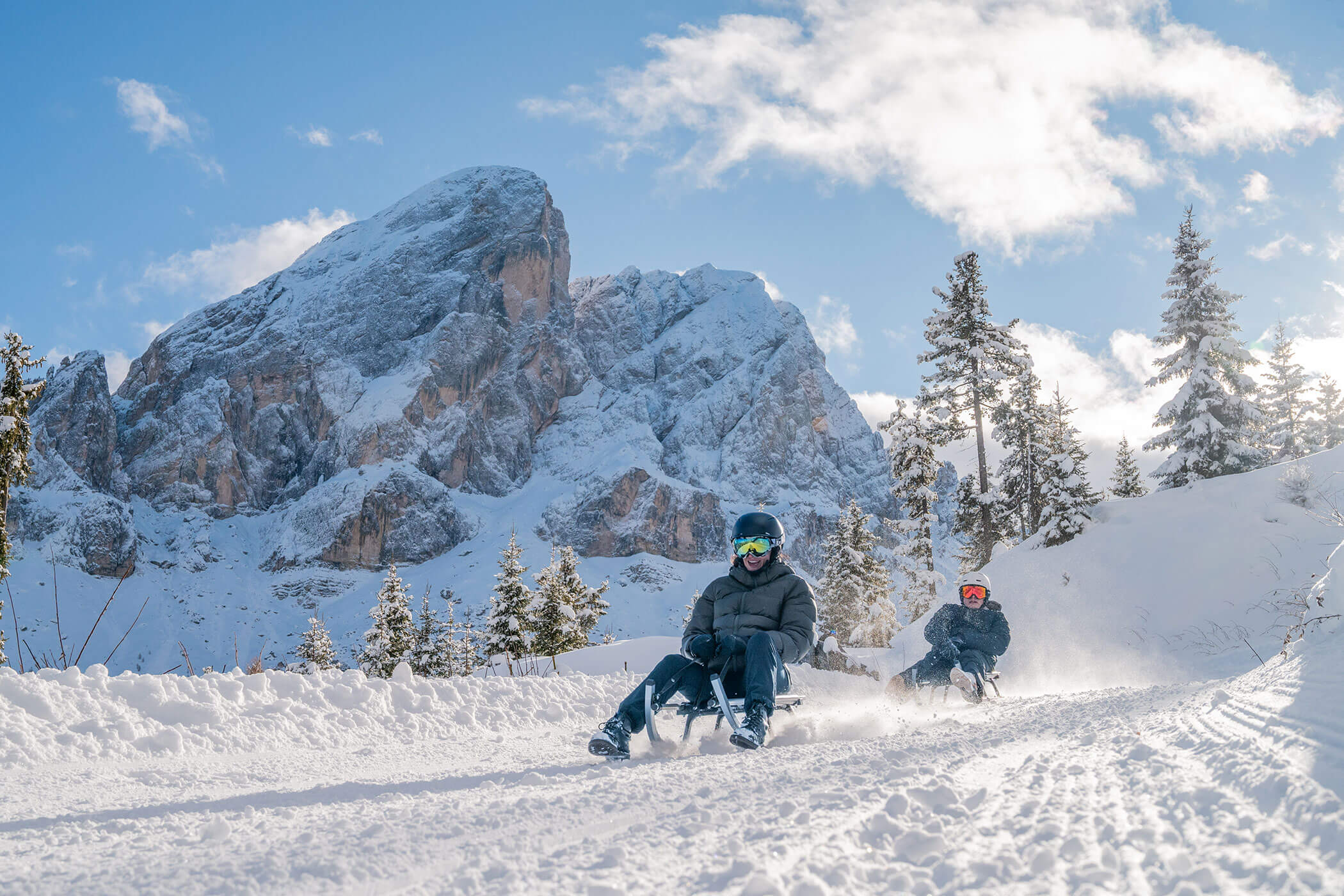 Zwei Menschen fahren mit dem Schlitten einen verschneiten Weg hinunter, im Hintergrund sind Berge und Kiefern zu sehen, über ihnen erstreckt sich ein blauer Himmel. - Hotel Oberwiesen