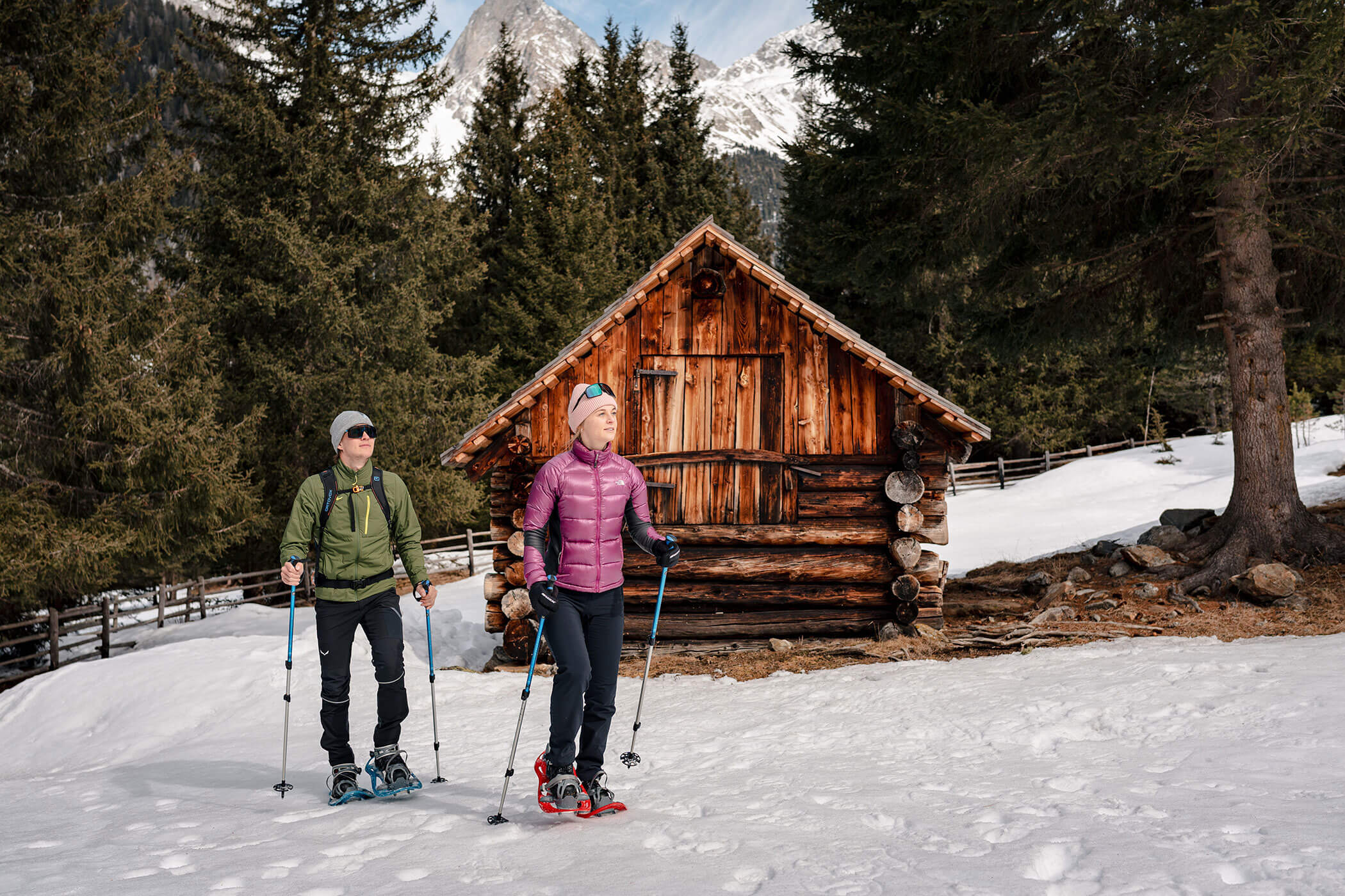 Zwei Menschen wandern mit Schneeschuhen vor einer Holzhütte, die von verschneiten Bäumen und Bergen umgeben ist. - Hotel Oberwiesen