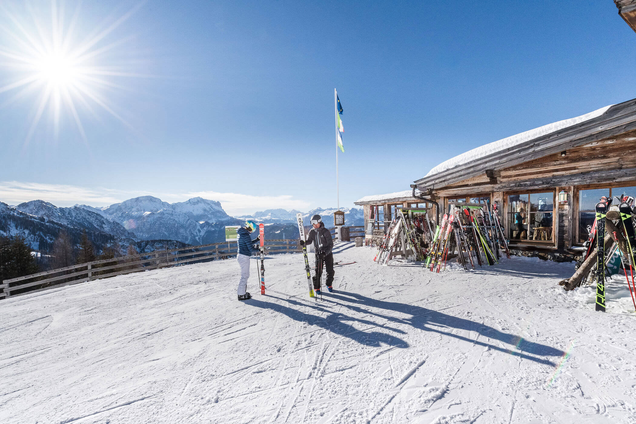 Zwei Skifahrer stehen vor einer verschneiten Berghütte im strahlenden Sonnenschein, im Hintergrund sind Skier und Berge zu sehen. - Hotel Oberwiesen