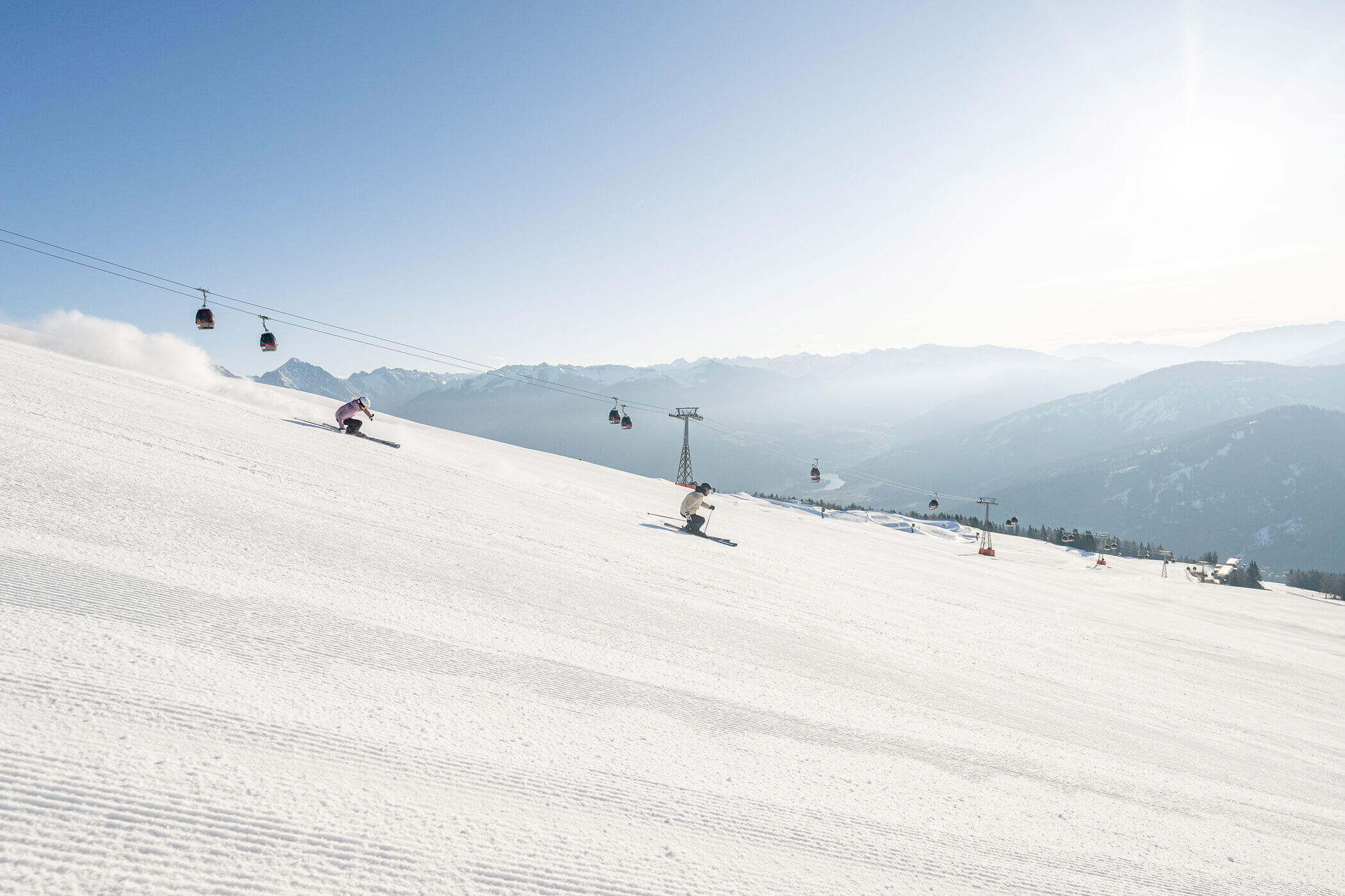 Skifahrer fahren einen verschneiten Berghang hinunter, im Hintergrund sind Skilifte und Berge zu sehen, während über ihnen ein strahlender Himmel zu sehen ist. - Hotel Oberwiesen