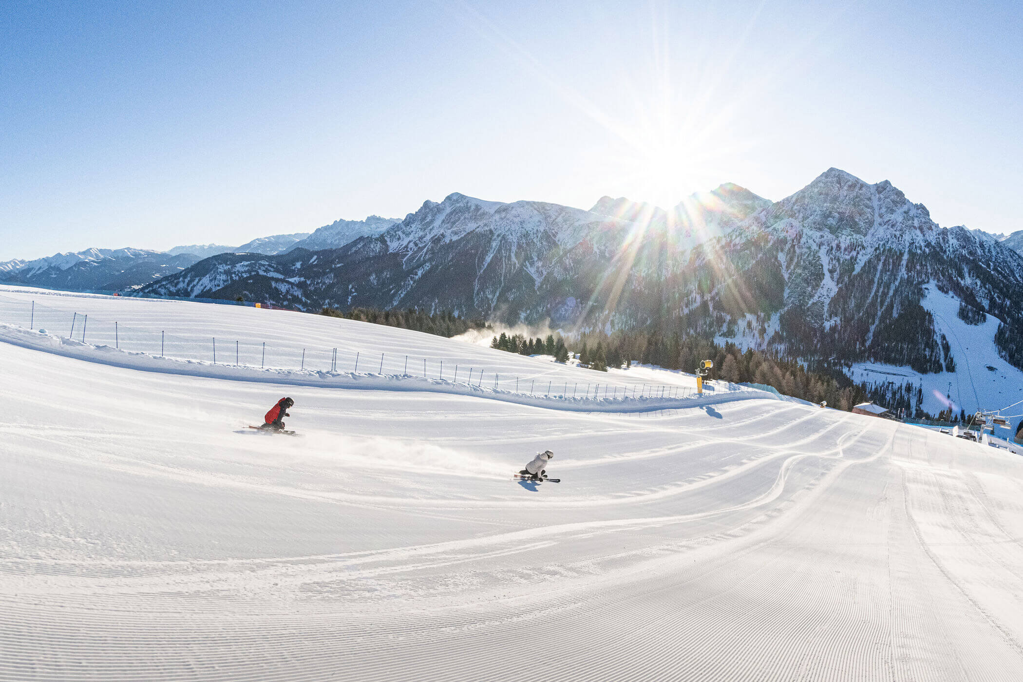 Zwei Skifahrer sausen einen präparierten, verschneiten Hang hinunter, im Hintergrund sind Berge und strahlender Sonnenschein zu sehen. - Hotel Oberwiesen