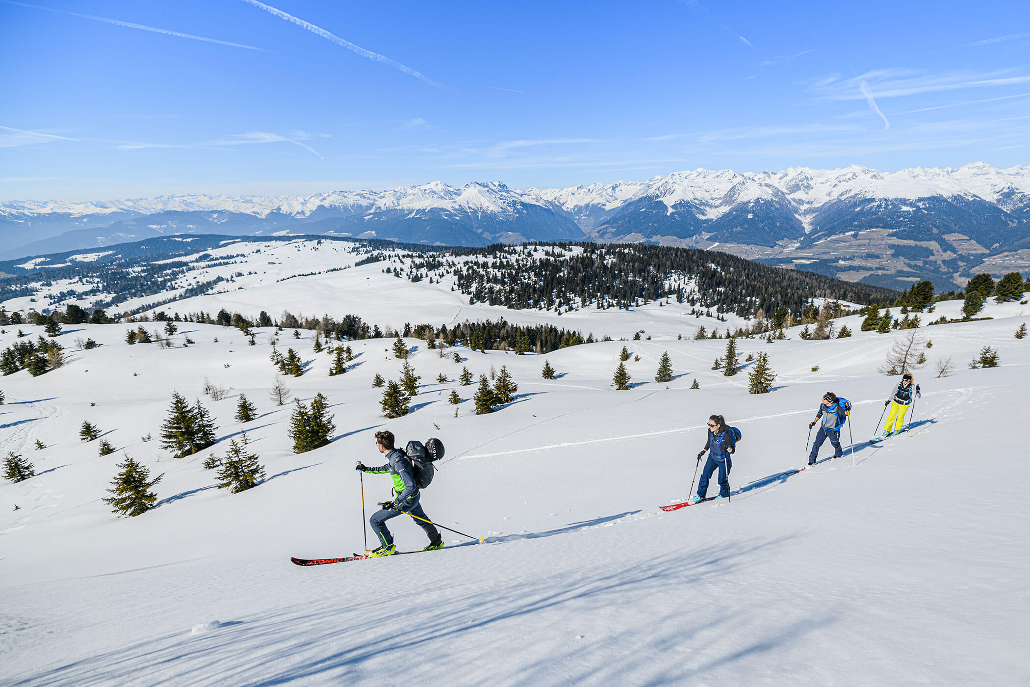 Vier Personen fahren auf Skiern einen verschneiten Berghang hinauf, im Hintergrund sind ferne Gipfel und ein blauer Himmel zu sehen. - Hotel Oberwiesen