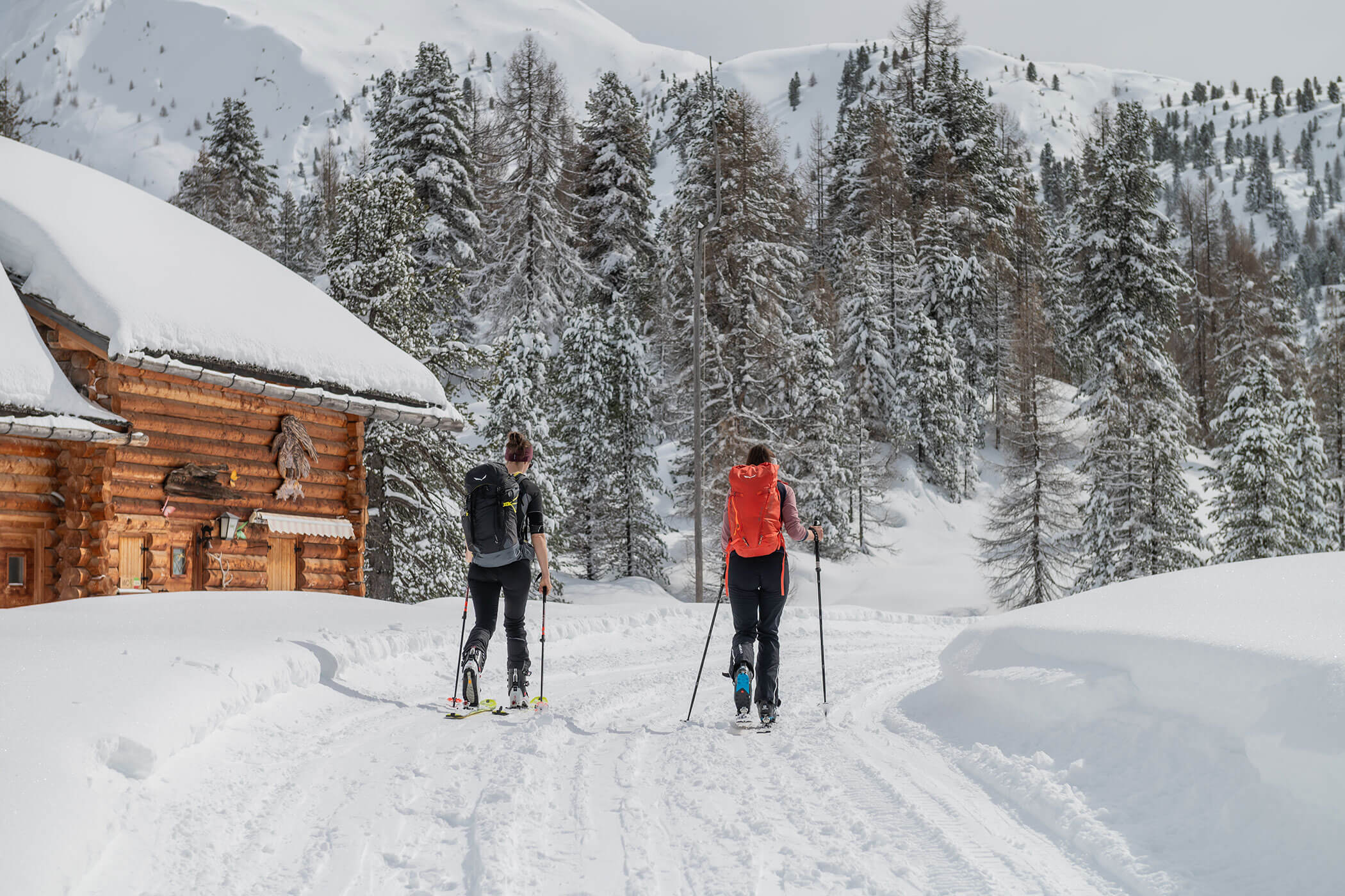 Zwei Menschen fahren mit Langlaufskiern in der Nähe einer Holzhütte in einem verschneiten Wald, im Hintergrund sind Berge zu sehen. - Hotel Oberwiesen