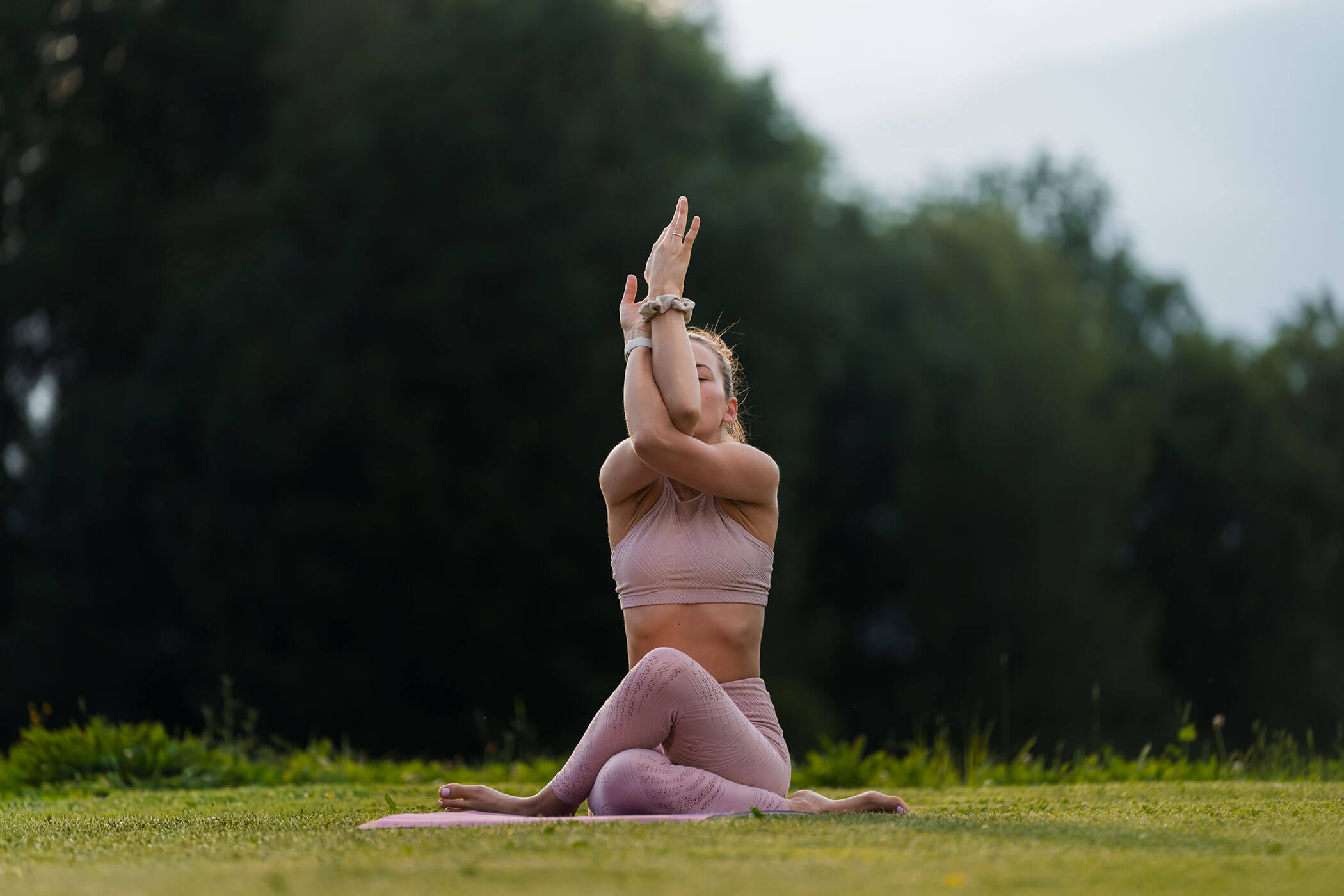 Eine Frau, die im Freien Yoga macht; sie sitzt auf einer Matte im Schneidersitz, die Arme sind erhoben und ineinander verschränkt. - Hotel Oberwiesen