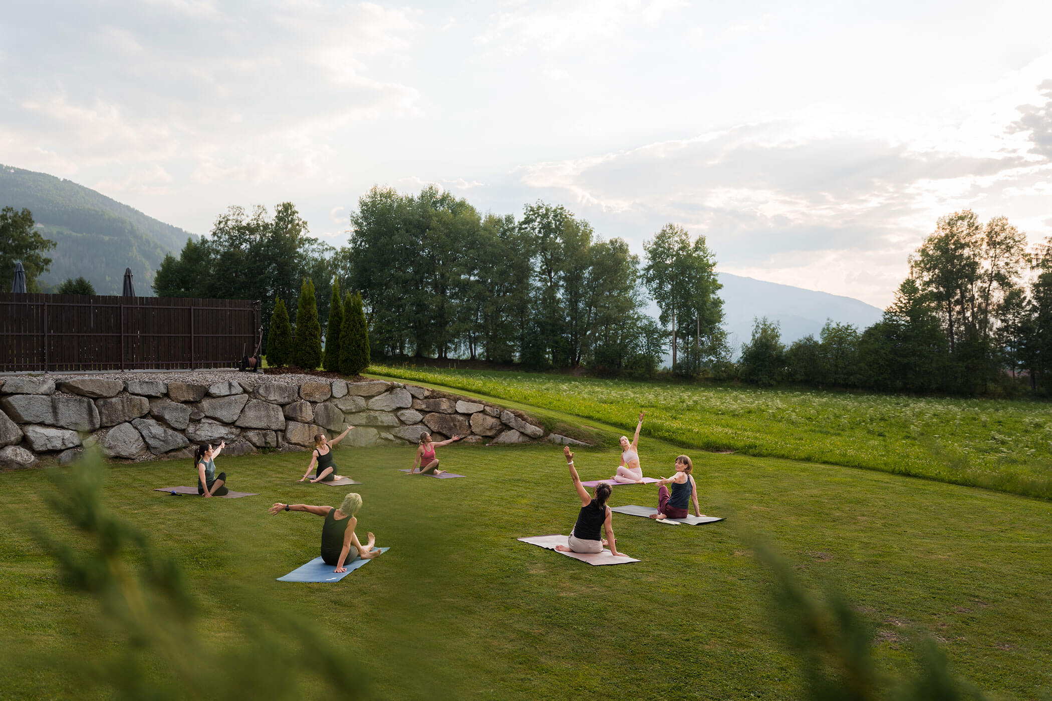 Eine Gruppe von Menschen macht Yoga auf Matten im Freien auf einer Rasenfläche, umgeben von Bäumen und Hügeln. - Hotel Oberwiesen