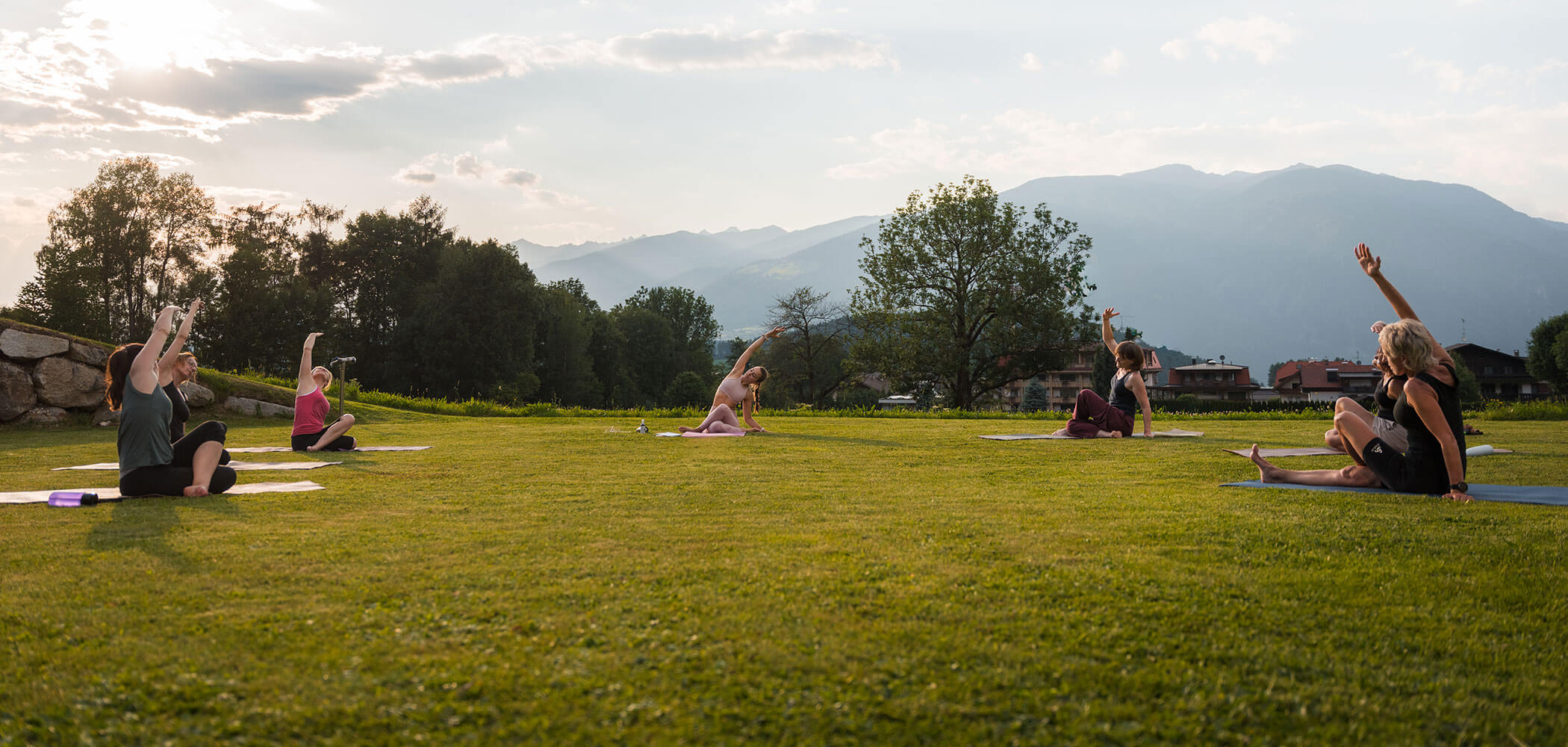 Fünf Menschen machen bei Sonnenuntergang auf Matten auf einer Wiese Yoga, im Hintergrund sind Bäume und Berge zu sehen. - Hotel Oberwiesen