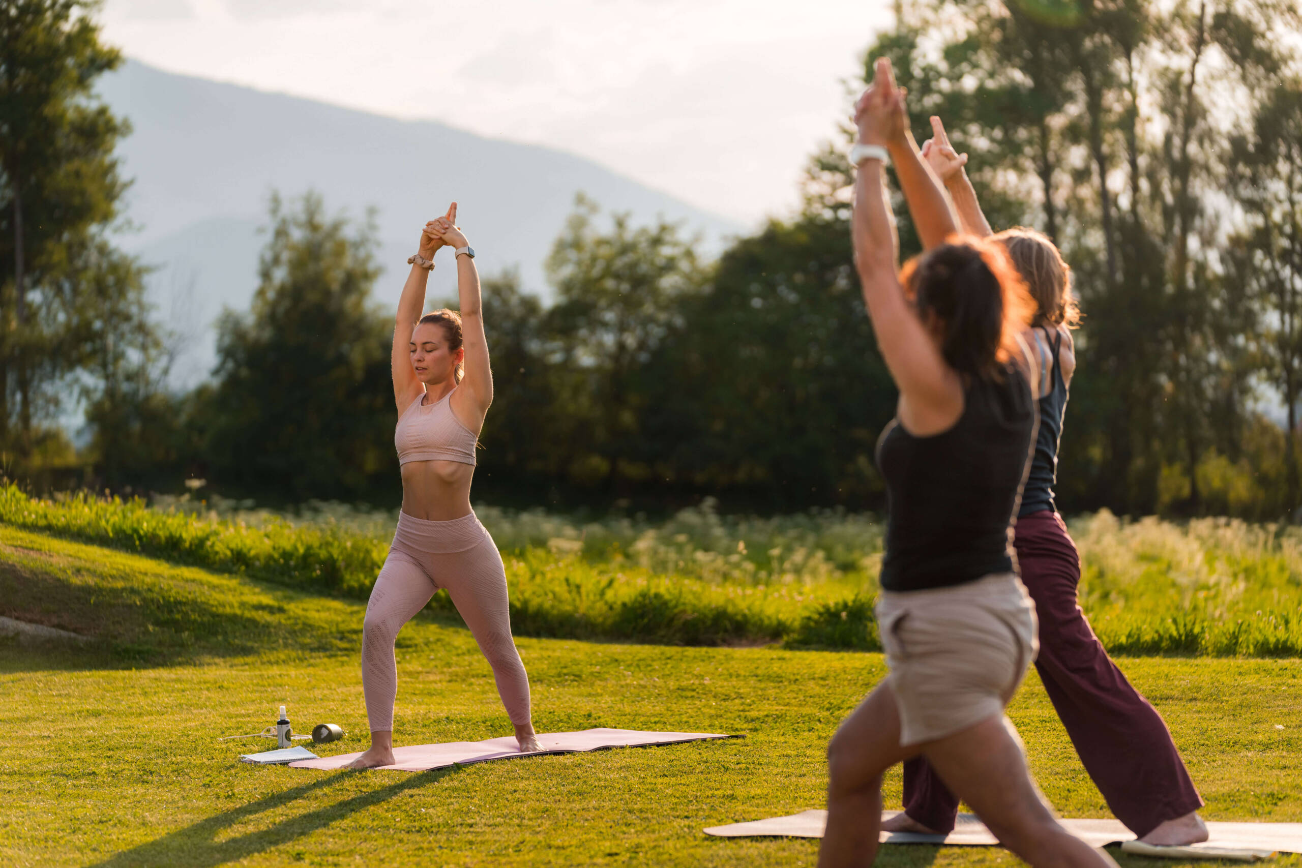 Drei Menschen, die draußen auf dem Rasen Yoga machen, die Arme erhoben, im Hintergrund Bäume und Berge. - Hotel Oberwiesen