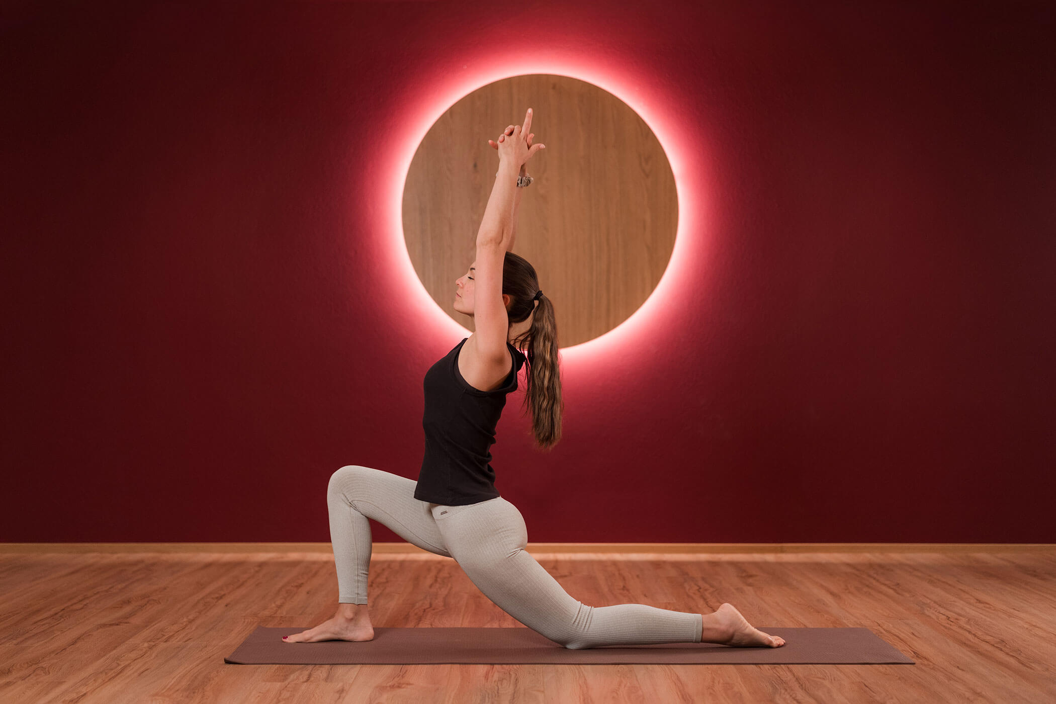 Eine Frau, die auf einer Matte Yoga macht und sich in einer tiefen Ausfallschritt-Position befindet, hinter ihr ist ein leuchtender Kreis an der roten Wand zu sehen. - Hotel Oberwiesen