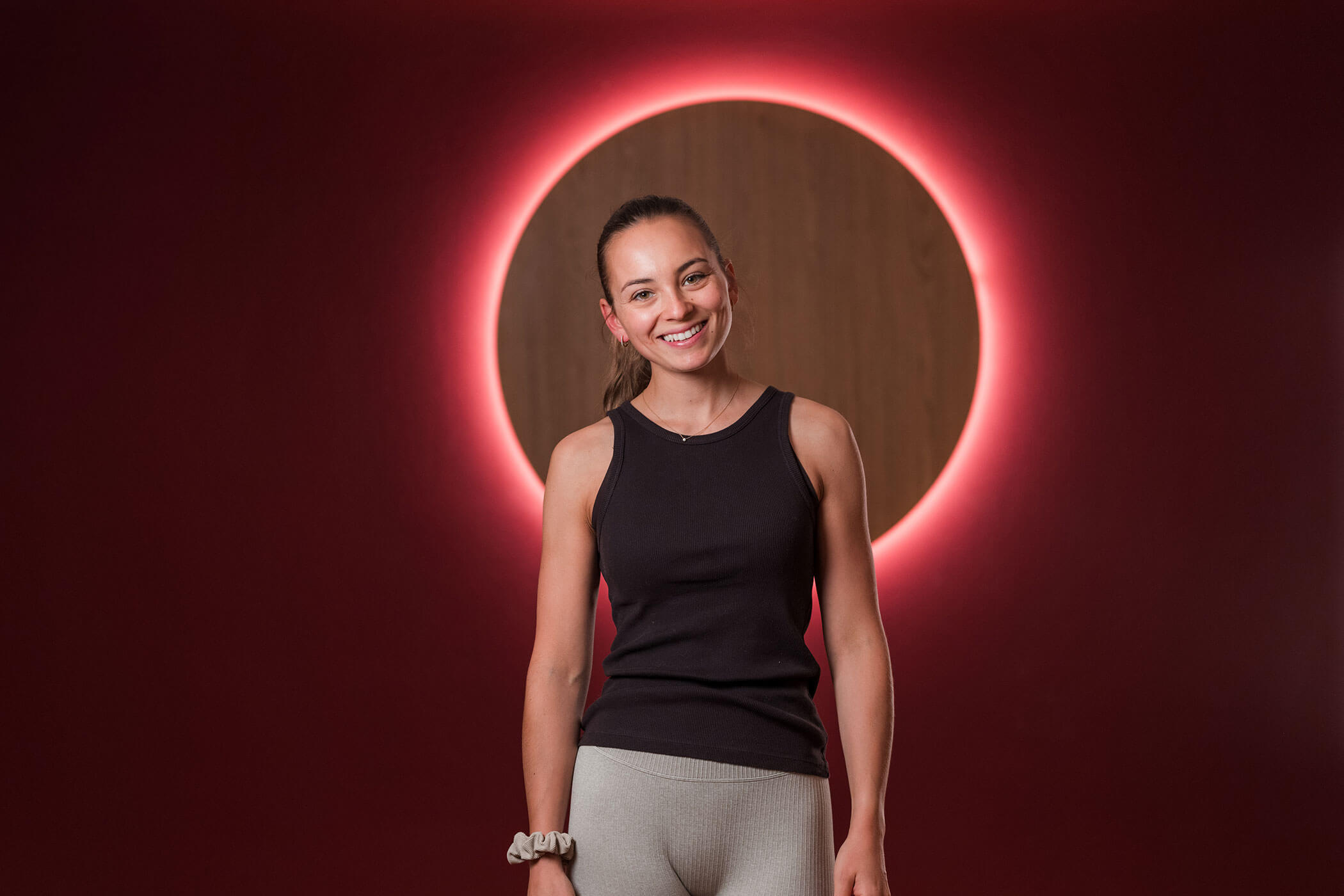 Eine Frau in Sportkleidung lächelt, während sie vor einer rot beleuchteten, runden Wandinstallation steht. - Hotel Oberwiesen