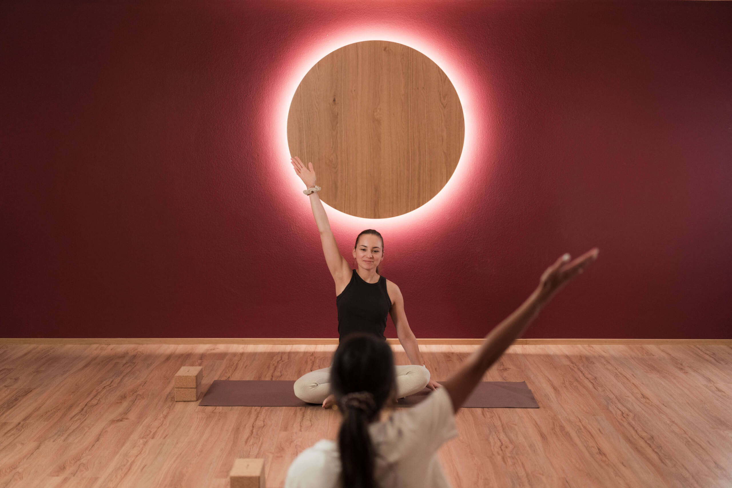 Zwei Menschen machen drinnen Yoga; einer sitzt im Schneidersitz und hebt einen Arm, an der Wand ist ein leuchtender Kreis zu sehen. - Hotel Oberwiesen